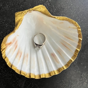 May include: A white seashell with a gold rim, used as a ring holder. A diamond ring is placed inside the shell.