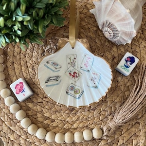 Puede incluir: Un adorno de concha blanca con ribete dorado, pintado con im&aacute;genes de fichas de mahjong. El adorno cuelga de una cuerda dorada y se muestra sobre una superficie tejida con cuentas de madera y una concha.