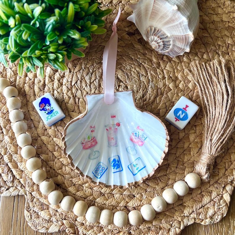 May include: A white seashell-shaped ornament with a pink ribbon for hanging. The shell is decorated with watercolor illustrations of cocktails and playing cards. Two game tiles and a wooden bead garland are also present.