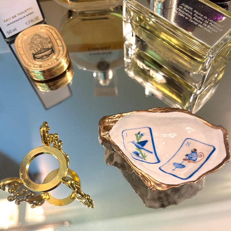 Op de afbeelding: Een opstelling van luxe items op een reflecterend oppervlak. Bevat een goudomrande oesterschelp schaal met twee ge&iuml;llustreerde kaarten, parfumflesjes en een gouden vergrootglas. De schaal heeft blauwe en witte illustraties.