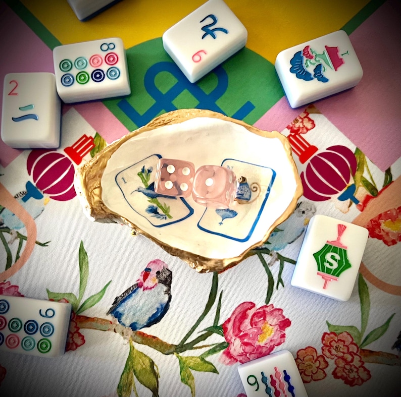 Op de afbeelding: Een verzameling witte Mahjong-tegels met kleurrijke markeringen en een paar roze dobbelstenen zijn gerangschikt rond een decoratieve oesterschelp. De schelp ligt op een stof met bloemenpatroon met roze, groene en blauwe accenten. De speelstukken zijn klaar om te spelen.