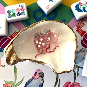 May include: A decorative oyster shell dish holds two pink dice with white dots and a small illustration. The shell is surrounded by Mahjong tiles on a colorful patterned surface. The shell has a gold-painted edge.