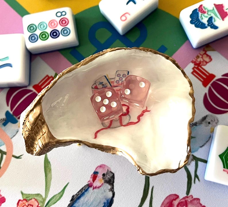 May include: A decorative oyster shell dish holds two pink dice with white dots and a small illustration. The shell is surrounded by Mahjong tiles on a colorful patterned surface. The shell has a gold-painted edge.