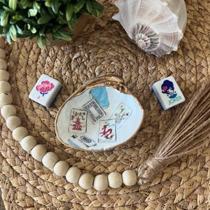May include: A decorative seashell dish with gold trim, holding several small, illustrated cards. Two Mahjong tiles are placed nearby. The scene is set on a woven mat, with a strand of wooden beads and a bundle of twine.