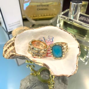 May include: A gold-colored seashell jewelry dish holds two rings. One ring is gold with clear stones, and the other has a large, square, light blue stone. The dish is white with gold trim.