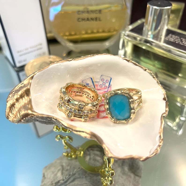 May include: A gold-colored seashell jewelry dish holds two rings. One ring is gold with clear stones, and the other has a large, square, light blue stone. The dish is white with gold trim.