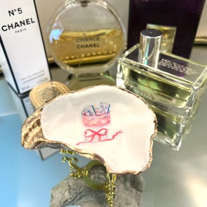 May include: An oyster shell trinket dish with a gold rim and a watercolor illustration of a pink box with playing cards. The dish is displayed with perfume bottles, including Chanel No. 5 and Chance Chanel.