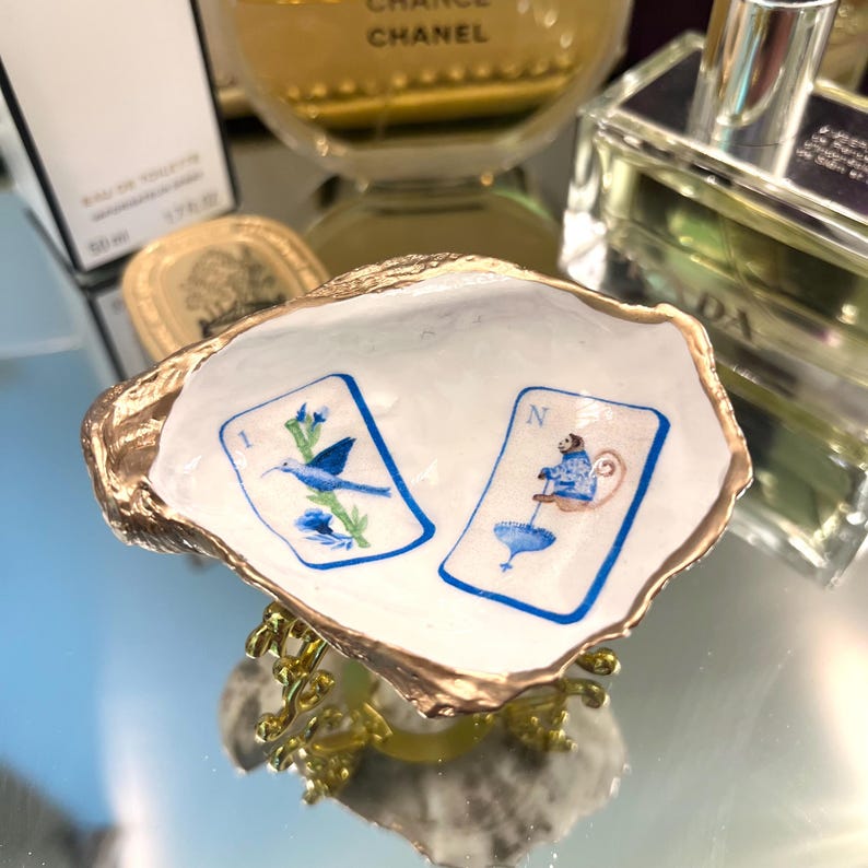 Op de afbeelding: Een decoratief oesterschelp sieradenschaaltje met een goudkleurige rand. De witte binnenkant heeft twee illustraties in speelkaartenstijl met blauwe en witte ontwerpen. Het schaaltje wordt ondersteund door een goudkleurige metalen standaard.