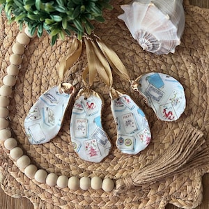 May include: Four oyster shell ornaments with gold trim and a playing card design. Each ornament has a different card design, including a Joker, a Queen of Hearts, and two other cards. The ornaments are hanging from gold ribbon and have a brown tassel.