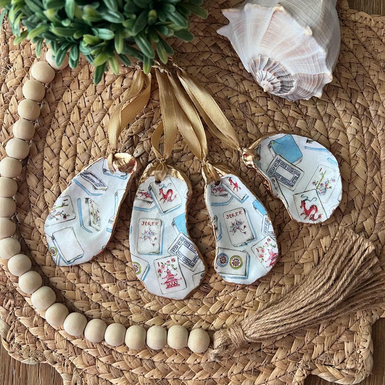 May include: Four oyster shell ornaments with gold trim and a playing card design. Each ornament has a different card design, including a Joker, a Queen of Hearts, and two other cards. The ornaments are hanging from gold ribbon and have a brown tassel.