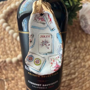 May include: A white ceramic ornament shaped like a seashell with a gold ribbon. The ornament is decorated with a colorful illustration of playing cards, including a Joker card. The text "JOKER" is visible on the card.
