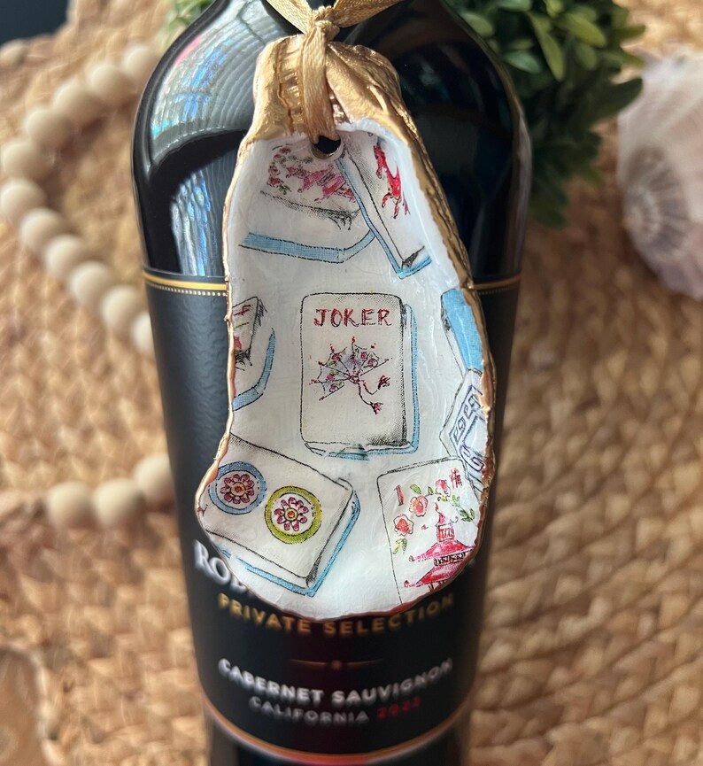 May include: A white ceramic ornament shaped like a seashell with a gold ribbon. The ornament is decorated with a colorful illustration of playing cards, including a Joker card. The text "JOKER" is visible on the card.