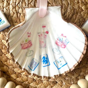 May include: A hand-painted seashell ornament with a pink ribbon for hanging. The white shell features watercolor illustrations of cocktails and playing cards. The shell has a gold-colored edge and is set against a woven background.