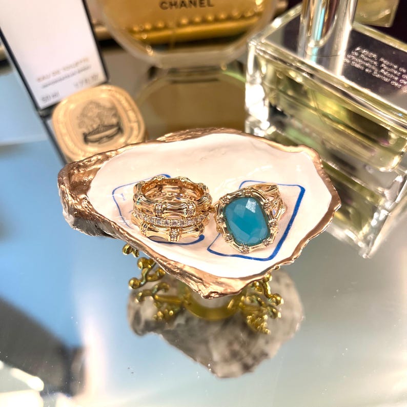 Op de afbeelding: Een decoratief sieradenbakje in de vorm van een oesterschelp met gouden rand, met twee ringen. E&eacute;n ring is goudkleurig met heldere stenen, de andere heeft een grote, rechthoekige, lichtblauwe steen. Het bakje staat op een goudkleurige, sierlijke standaard.