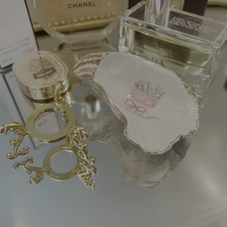 May include: A decorative oyster shell dish with a gold-painted rim and a painted pink baby carriage design. A gold-colored decorative stand and perfume bottles are also visible.