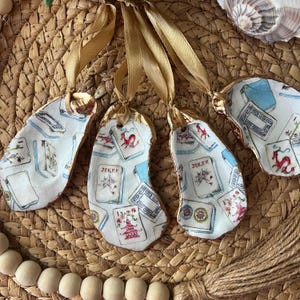 May include: Four white oyster shell ornaments with gold trim and a blue and white playing card design. The ornaments are hanging from gold ribbon.