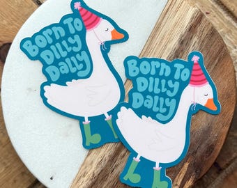May include: Two stickers featuring a cartoon goose wearing a party hat and teal boots. The stickers have the text "Born To Dilly Dally" in a teal bubble. The stickers are on a white and wood grain coaster.
