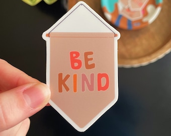 May include: A sticker in the shape of a banner with the words "BE KIND" in colorful letters. The banner is a light brown color with a white top. The sticker is held by a hand.