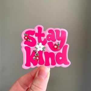 May include: A pink sticker with the words "Stay Kind" in a bubble font. The sticker has a light pink border and is decorated with small floral accents in red, white, and green.