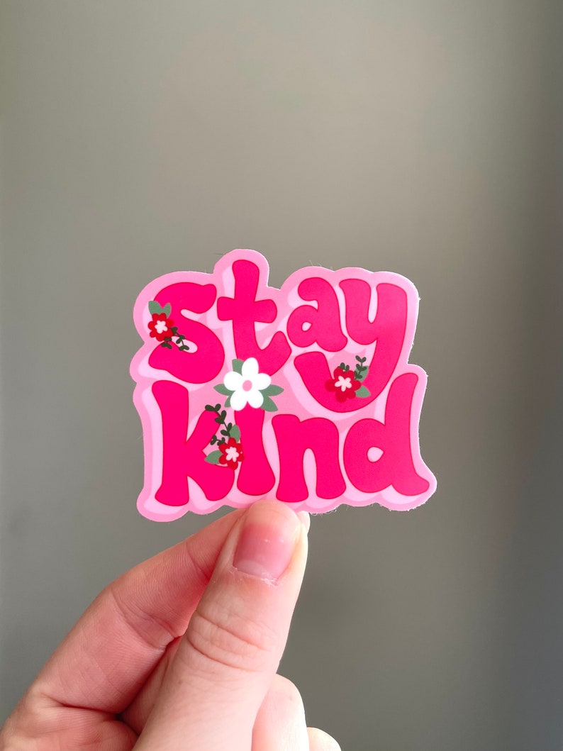 May include: A pink sticker with the words "Stay Kind" in a bubble font. The sticker has a light pink border and is decorated with small floral accents in red, white, and green.