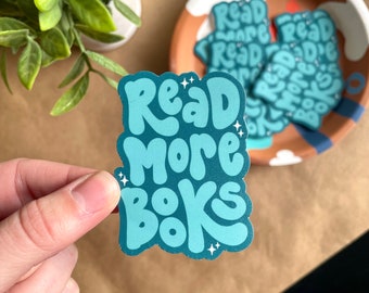 Może przedstawiać: Turkusowa naklejka z napisem "Read More Books" w stylu retro. Naklejka jest trzymana w dłoni, z dodatkowymi naklejkami w dekoracyjnej misce. Tło to brązowa powierzchnia z małą rośliną.