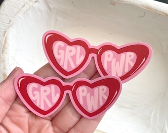 Op de afbeelding: Twee hartvormige stickers met rode frames en roze centra. De stickers lezen "GRL PWR" in een gestileerd lettertype. De stickers staan op een lichtroze achtergrond.