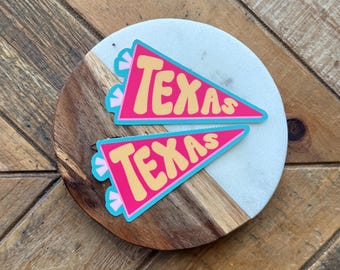 May include: Two pink pennant-shaped stickers with the word "TEXAS" in yellow, outlined in blue. The stickers are on a white and brown wooden surface. The stickers have a retro design.