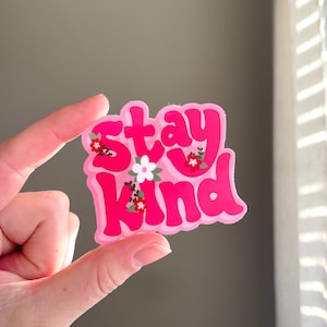 May include: A pink sticker with the words "Stay Kind" in a bubble font, decorated with small floral accents. The sticker is held between fingers against a neutral background.