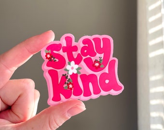 May include: A pink sticker with the words "Stay Kind" in a bubble font, decorated with small floral accents. The sticker is held between fingers against a neutral background.