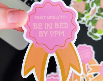 May include: A pink and orange sticker with the text "MOST LIKELY TO: BE IN BED BY 9PM". The sticker has a ribbon design and is held by a hand. Other stickers are in the background.
