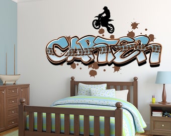 Op de afbeelding: Een bruin houten bedframe met een blauw-groen gestreept dekbed en een blauw-wit gepatroneerd kussen. De muur achter het bed heeft een bruin en blauwe graffiti-stijl sticker met de naam "Carter" en een silhouet van een persoon die een crossmotor rijdt.