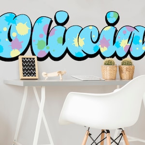 Puede incluir: Un diseño de salpicaduras de pintura azul y blanco con el nombre Olivia escrito en un contorno negro en negrita. El diseño está en una pared blanca sobre un escritorio con una silla blanca, un avión de juguete de madera y una pequeña imagen enmarcada en negro.