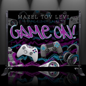 May include: A black and purple banner with the text "Mazel Tov Levi" and "Game On!" in a pink and blue font. The banner features a variety of video game controllers and headphones.