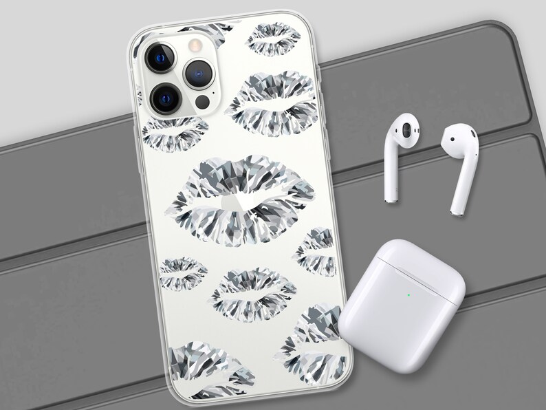 May include: A clear phone case with a white background and a repeating pattern of black and white diamond-shaped lips. The case is shown on a grey surface with a pair of white wireless earbuds and a white charging case.