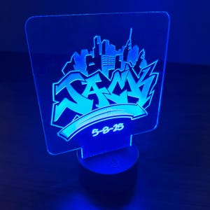 Puede incluir: Una luz nocturna de acrílico azul con una ciudad blanca y el nombre "JAMA" en letras de estilo graffiti. La fecha "5-8-25" también está incluida en el diseño.