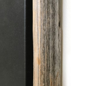May include: Close-up of the edge of a black canvas painting framed in a rustic wood frame. The wood has a natural, weathered finish with knots and grain patterns.