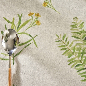 May include: A close-up of a linen tablecloth with a floral pattern featuring green ferns and yellow flowers. A silver knife and spoon with wooden handles are placed on the tablecloth.