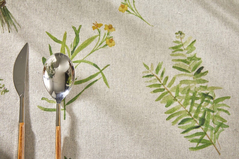 May include: A close-up of a linen tablecloth with a floral pattern featuring green ferns and yellow flowers. A silver knife and spoon with wooden handles are placed on the tablecloth.