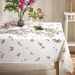 May include: White tablecloth with a green and red berry pattern. The tablecloth is laid on a wooden table with a vase of pink flowers and a vase of green berries on top.