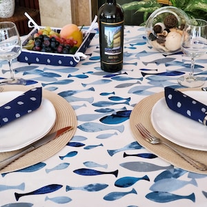 以下が含まれることがあります： 青い魚の模様の白いテーブルクロスが敷かれたダイニングテーブル。白いお皿、紺色のナプキン、ワイングラスがセットされています。果物のバスケットとワインボトルもテーブルにあります。