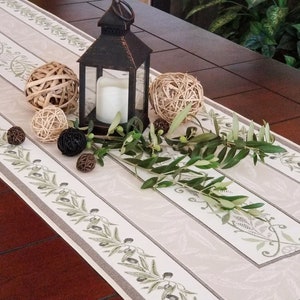 May include: A white table runner with a gray border and a repeating olive branch design. The runner is decorated with a black lantern, a sprig of greenery, and several woven balls.