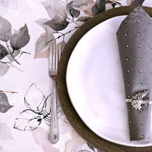 May include: A formal table setting featuring a white plate atop a wooden charger, with a gray napkin adorned with rhinestones and a decorative napkin ring. A silver fork rests on a floral patterned tablecloth.