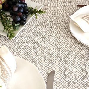 May include: A white tablecloth with a brown geometric pattern. Two place settings are shown with white plates, white napkins with brown trim, and silverware. A plate of grapes and rosemary is in the upper left corner.