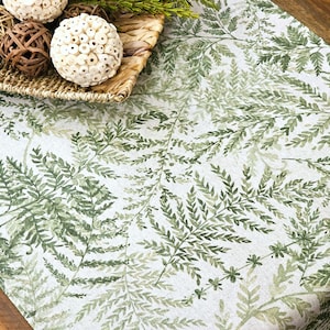 May include: A white table runner with a green fern leaf pattern. The runner is decorated with a woven basket containing decorative spheres and greenery, creating a natural, botanical theme.
