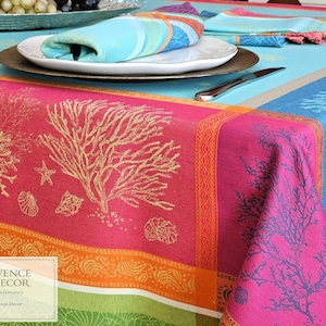 May include: A colorful tablecloth with a coral reef design. The tablecloth features a vibrant pink and orange border, with a turquoise and blue background. The design includes coral, starfish, and seashells. A plate with a napkin and silverware are on the table.