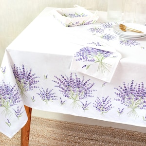 May include: White tablecloth with a lavender floral design. The tablecloth has a border of lavender bouquets and matching napkins. A basket and plates with gold utensils are on the table.