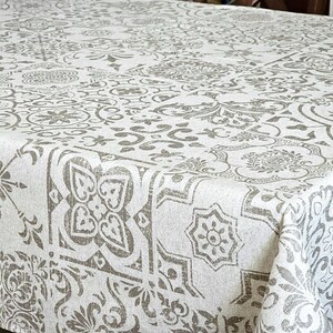 May include: A white tablecloth with a repeating pattern of gray floral and geometric designs. The fabric has a textured appearance, and the pattern is evenly distributed across the surface. The tablecloth is rectangular and appears to be on a dining table.