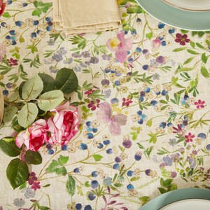 May include: A white tablecloth with a floral pattern featuring pink, purple, and blue flowers. The tablecloth is decorated with a vase of pink roses and a set of plates.