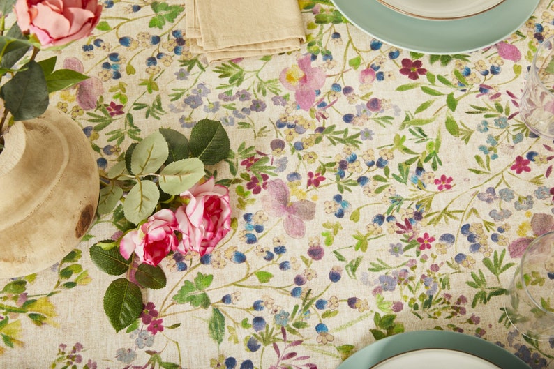 May include: A white tablecloth with a floral pattern featuring pink, purple, and blue flowers. The tablecloth is decorated with a vase of pink roses and a set of plates.