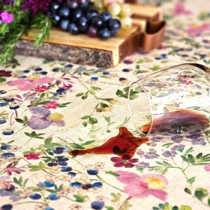 May include: A floral tablecloth with a spilled glass of red wine. The tablecloth features a pattern of pink, purple, and blue flowers. A wooden cutting board with grapes and a sprig of rosemary is in the background.